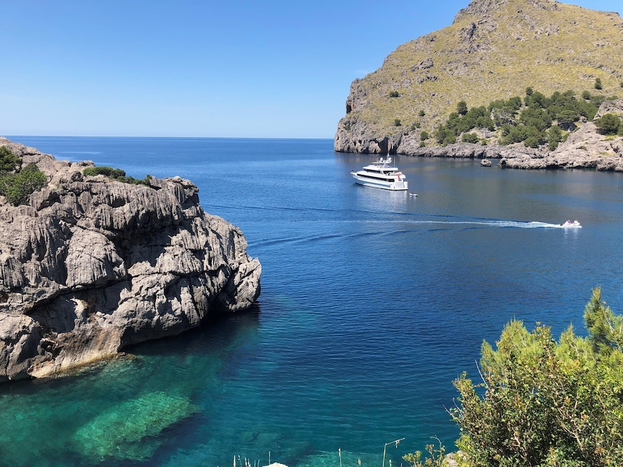 A yacht on a clear blue sea, rocky coastline, and a hill with sparse vegetation.