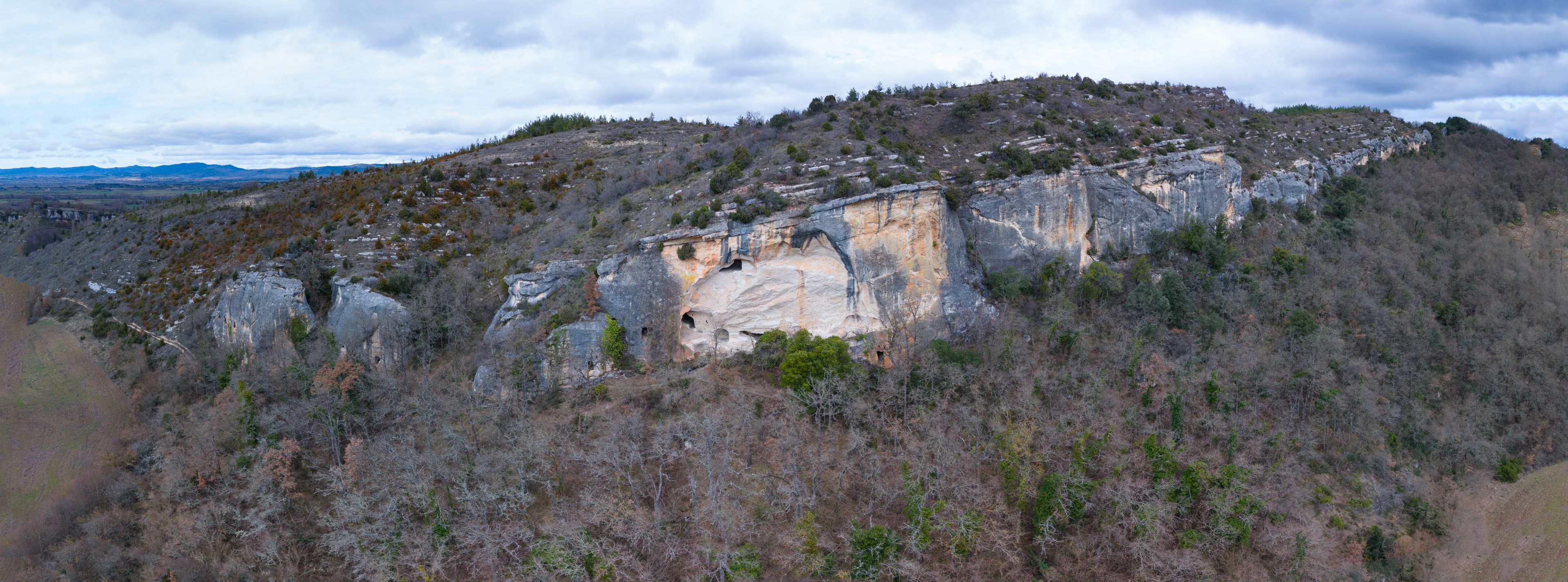 Un paysage montagneux accidenté, dominé par une formation rocheuse remarquable, avec des grottes et une forêt dense à ses pieds.