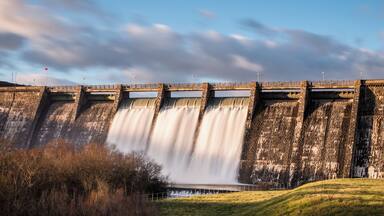 A large dam with multiple water outlets, a grassy foreground, and a partly cloudy sky.