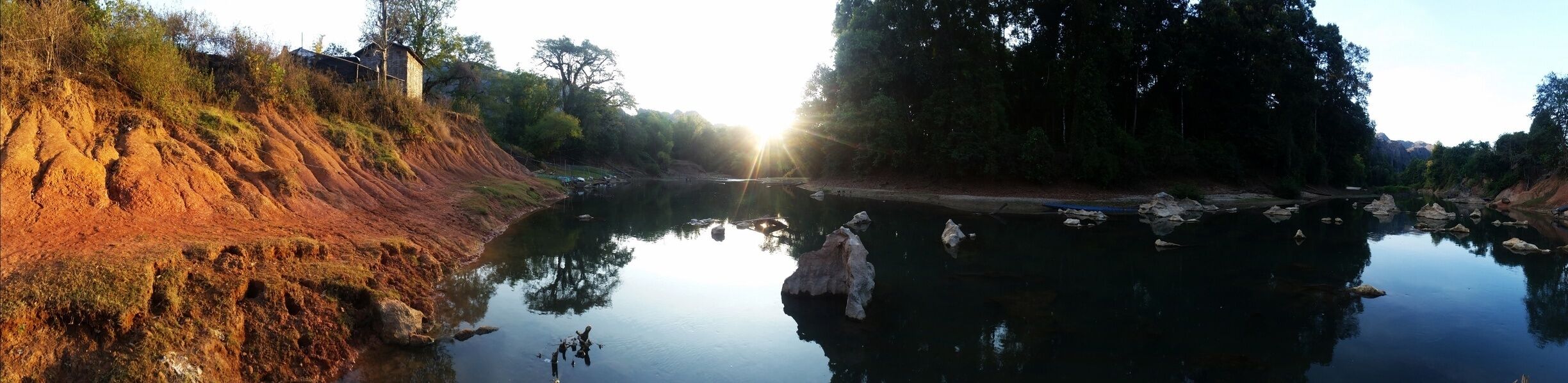 A calm river with reflections of trees and rocks, a dirt embankment, and a small building in the background.