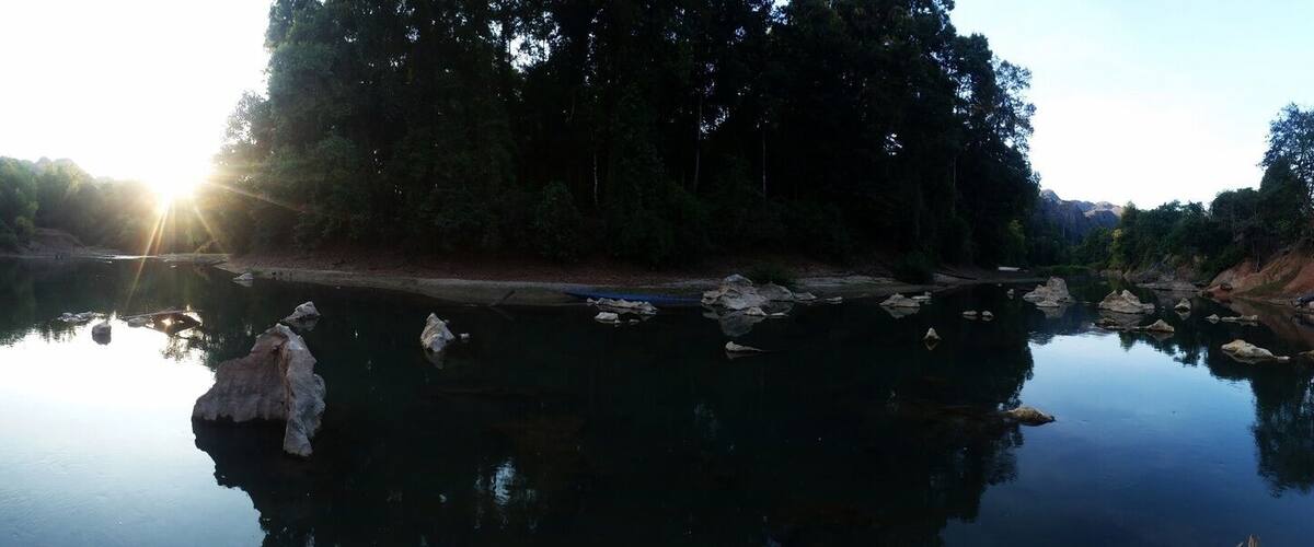 A calm river with reflections of trees and rocks, a dirt embankment, and a small building in the background.