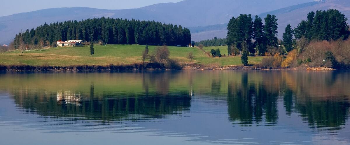 A calm lake with a house on the left, surrounded by trees and mountains in the background.