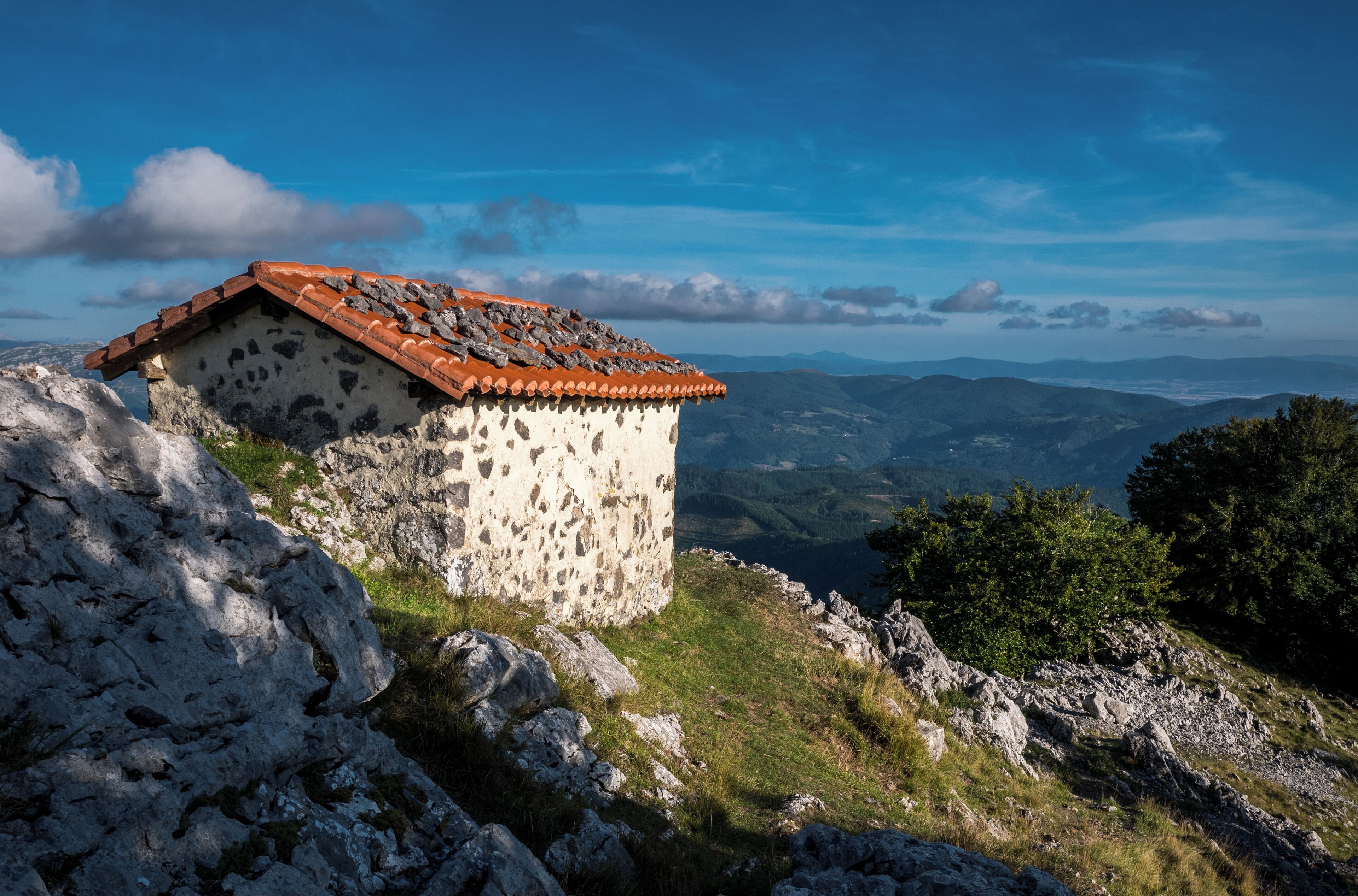 A stone building with a red-tiled roof on a rocky hillside overlooking a valley.