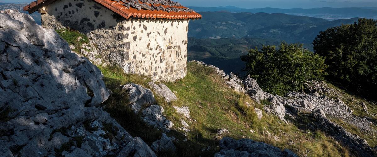 A stone building with a red-tiled roof on a rocky hillside overlooking a valley.