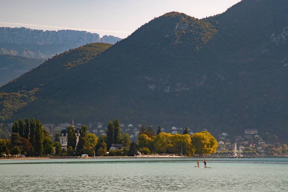 Ein See mit Bergen im Hintergrund, einigen Bäumen und einigen Gebäuden am Ufer.