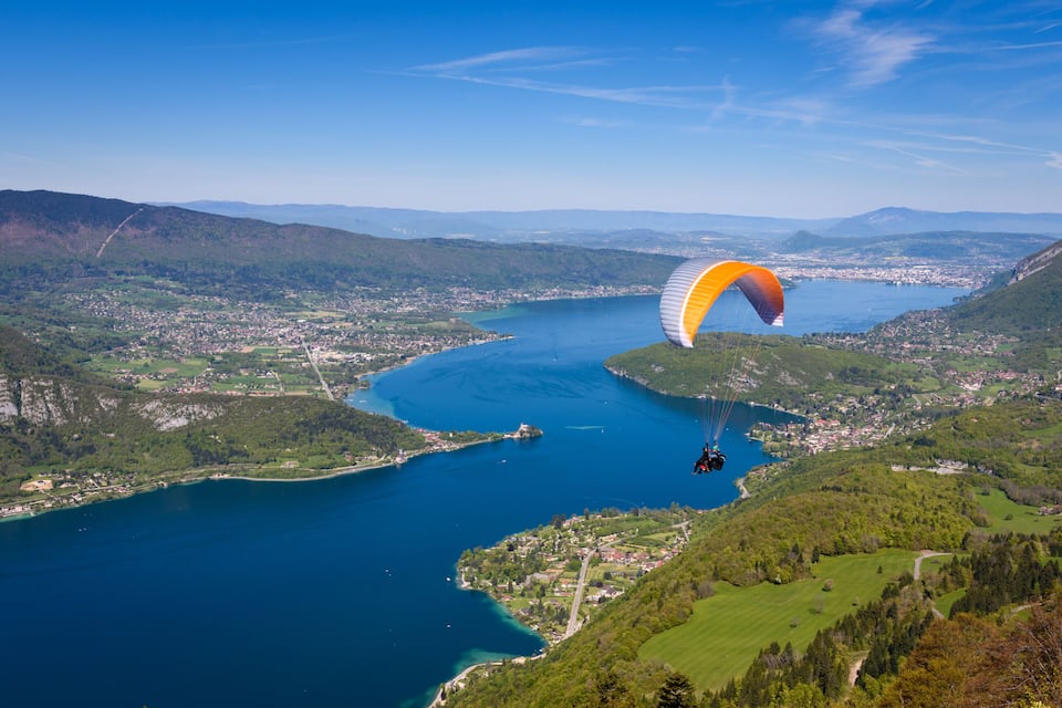 Una persona fa parapendio sopra una località costiera con un'ampia distesa d'acqua e colline circostanti.