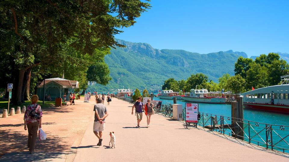 A promenade with people walking, a dog, and boats on a lake with mountains in the background.