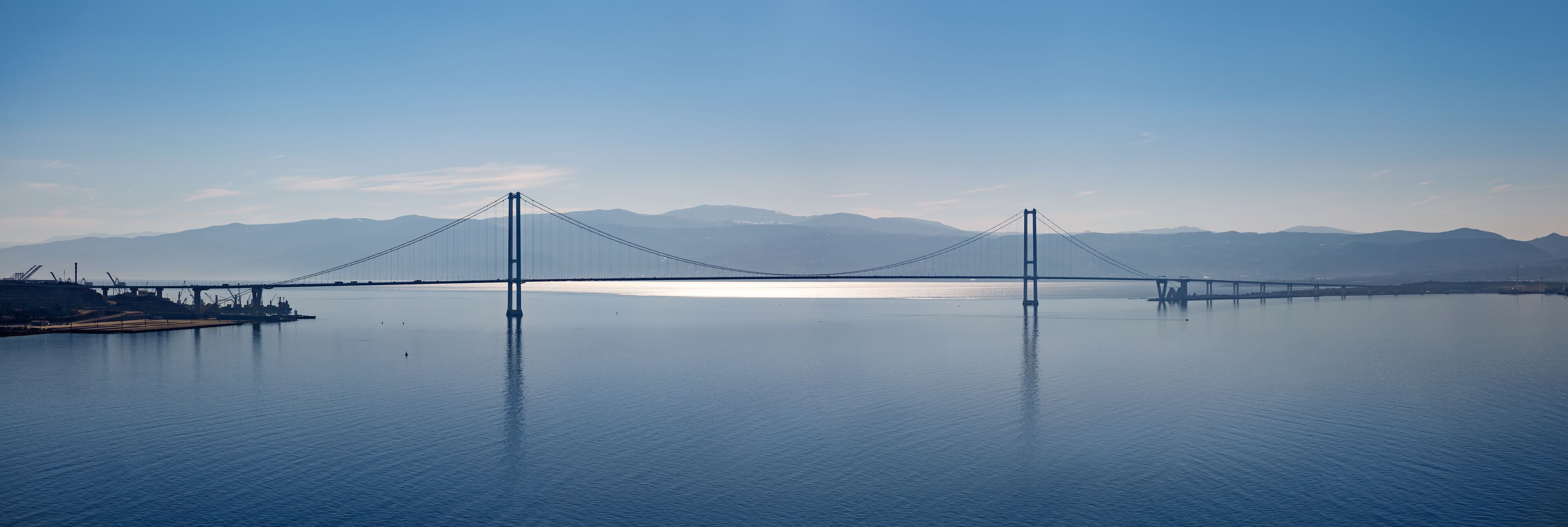 A suspension bridge over a calm body of water with mountains in the background.