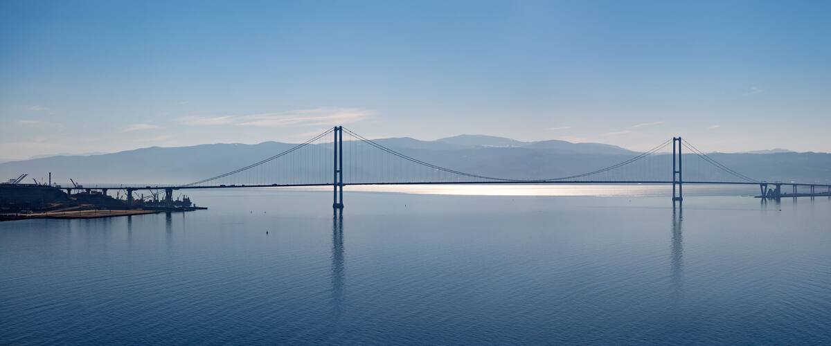 A suspension bridge over a calm body of water with mountains in the background.