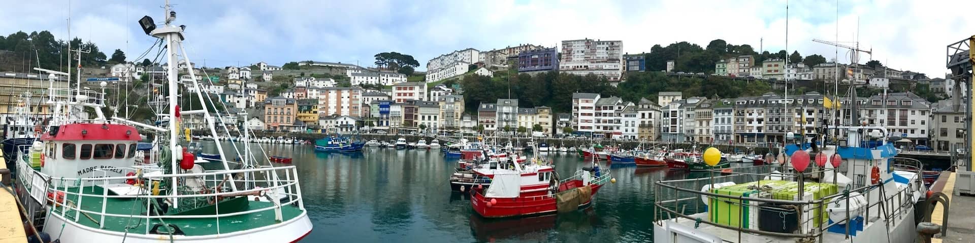 A harbor with multiple boats docked, a lighthouse, and a hillside with buildings.