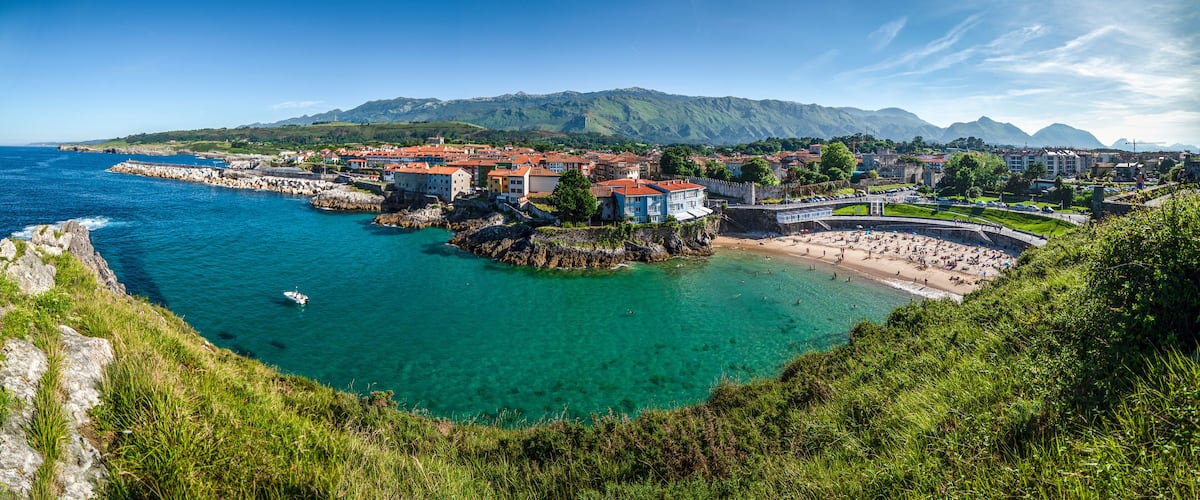 A coastal town with clear turquoise waters, a rocky shoreline, and a mountainous backdrop.
