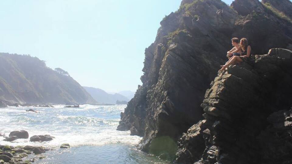 A rocky coastline with waves crashing against the shore and a couple sitting on a rock formation.