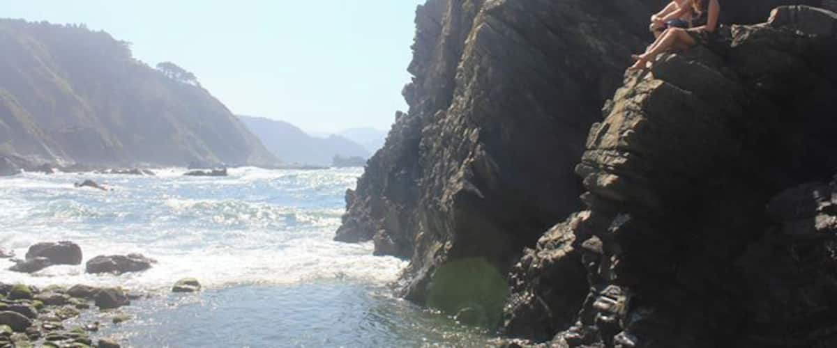 A rocky coastline with waves crashing against the shore and a couple sitting on a rock formation.