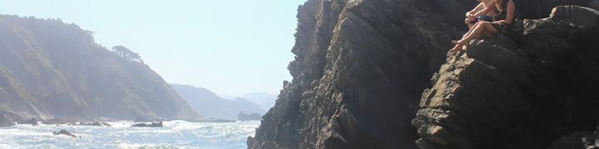 A rocky coastline with waves crashing against the shore and a couple sitting on a rock formation.