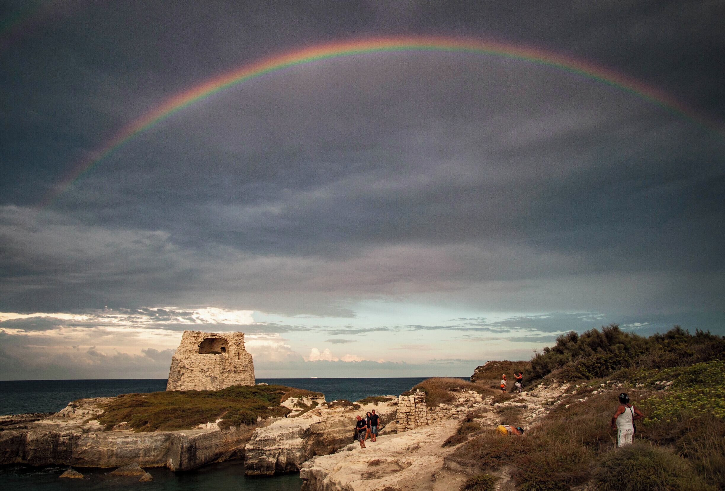 A coastal landscape with a rainbow, rocky cliffs, and a historical stone structure.