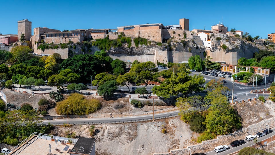 The image captures the medieval fortress in Cagliari's Castello district, surrounded by greenery, roads, and a mix of modern and traditional buildings.
