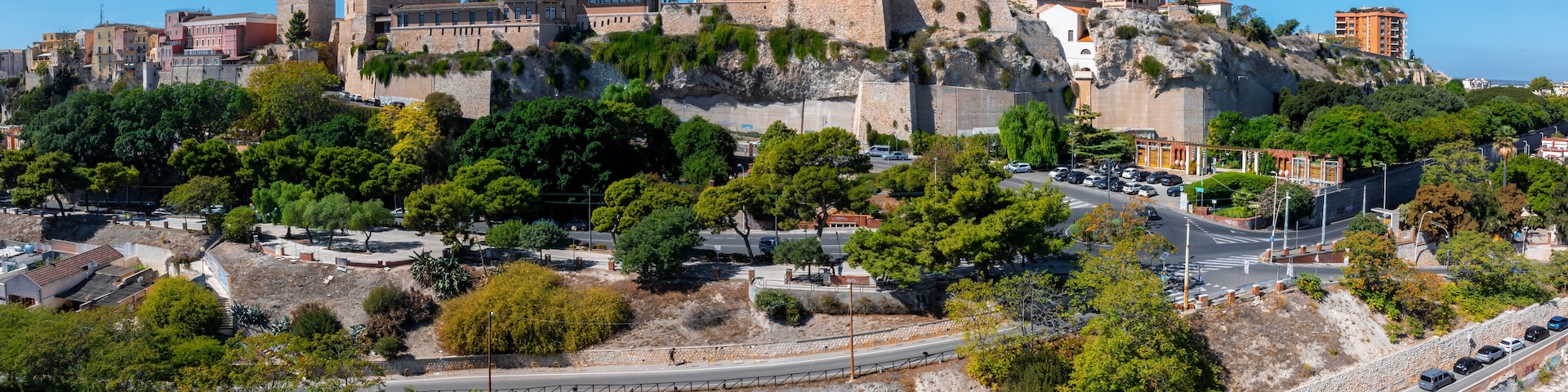 The image captures the medieval fortress in Cagliari's Castello district, surrounded by greenery, roads, and a mix of modern and traditional buildings.