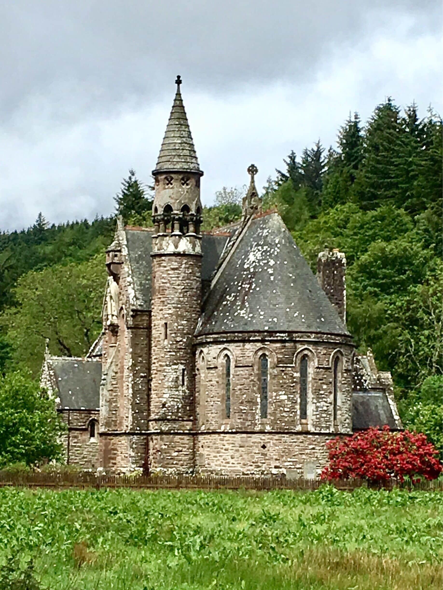 A historic stone church with a pointed tower and arched windows, surrounded by lush greenery and a red flowering bush.
