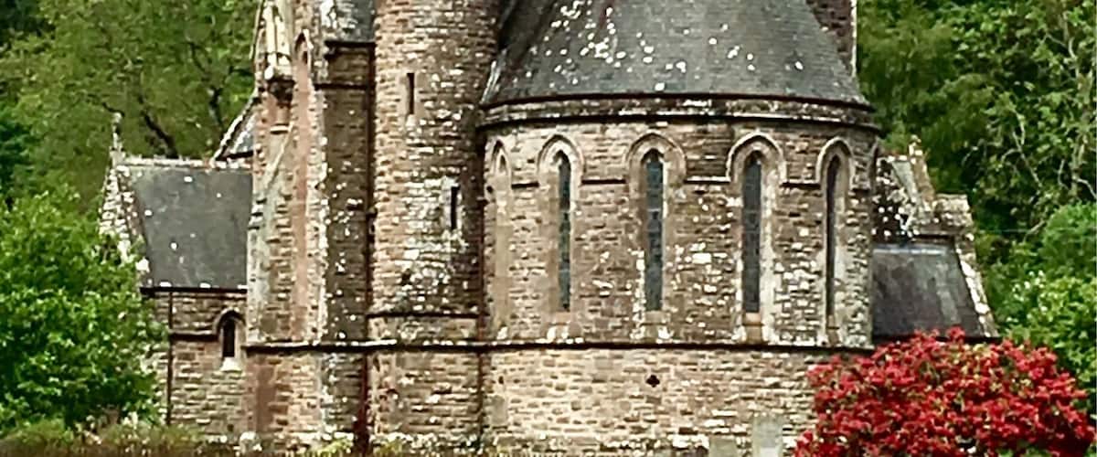 A historic stone church with a pointed tower and arched windows, surrounded by lush greenery and a red flowering bush.