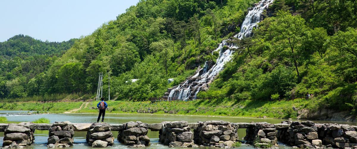 A waterfall cascading down a lush green hillside, a rocky bridge over a river, and a person standing on the bridge.