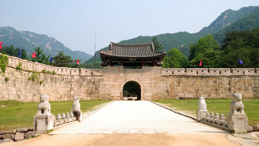 A traditional gate with a tiled roof, flanked by stone lion statues, leading to a mountainous landscape.