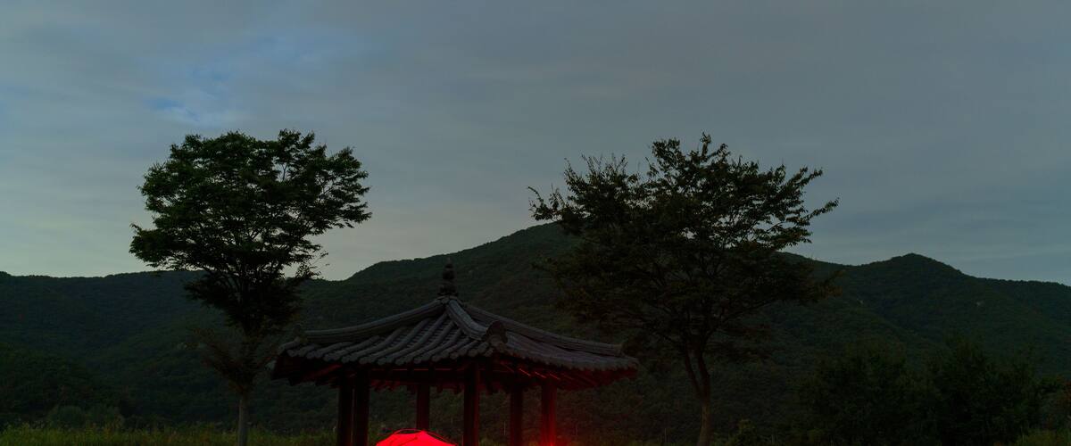 A traditional wooden pavilion with a red lantern in the foreground, set against a backdrop of a hilly landscape with trees.