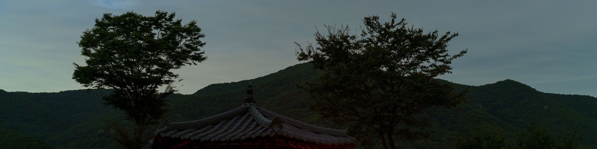 A traditional wooden pavilion with a red lantern in the foreground, set against a backdrop of a hilly landscape with trees.