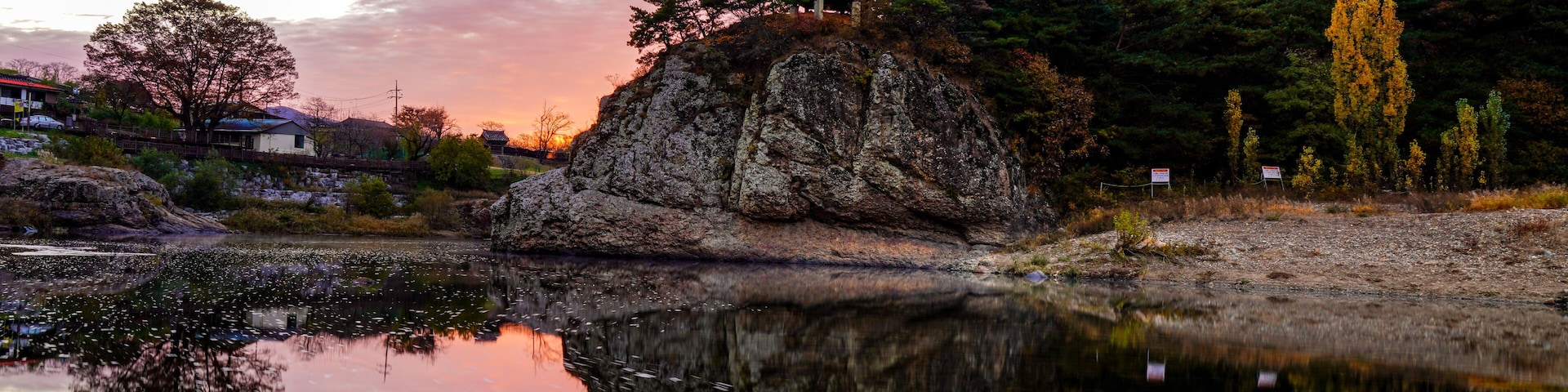 Una pagoda tradicional sobre un afloramiento rocoso junto a un estanque que refleja el cielo al atardecer, con árboles otoñales.