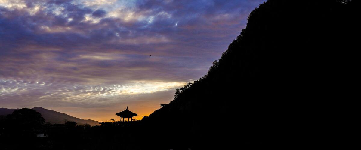 A sunset over a mountainous landscape with a traditional pavilion silhouetted against the sky.