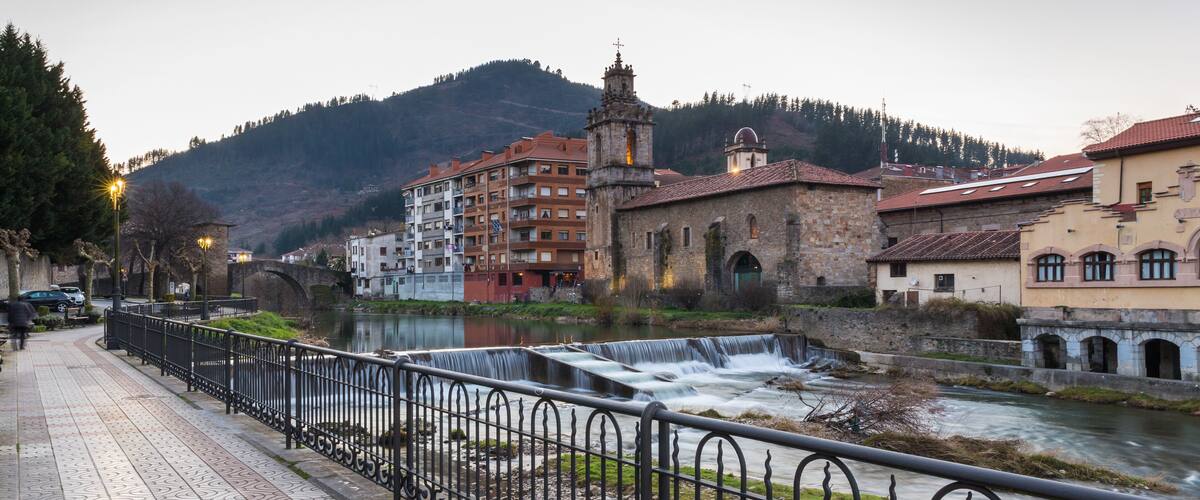 A riverside walkway with a stone bridge, a church, and residential buildings.