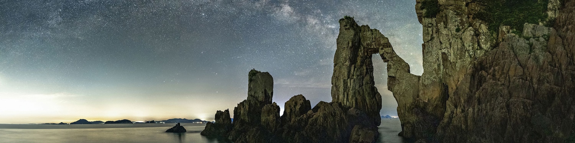 A starry night sky over a coastal landscape with rock formations and a calm sea.