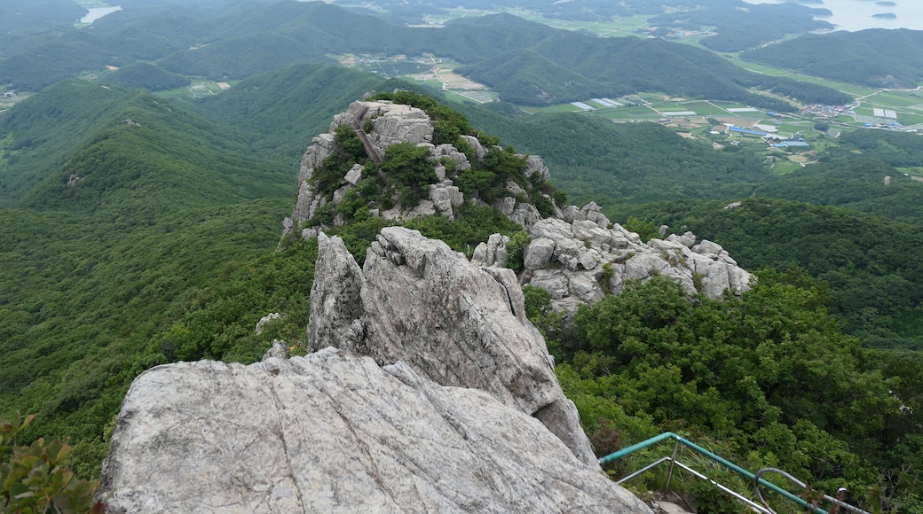 Un paysage montagneux avec un sommet rocheux imposant, un escalier menant au sommet, et une vallée avec des champs et une rivière au loin.