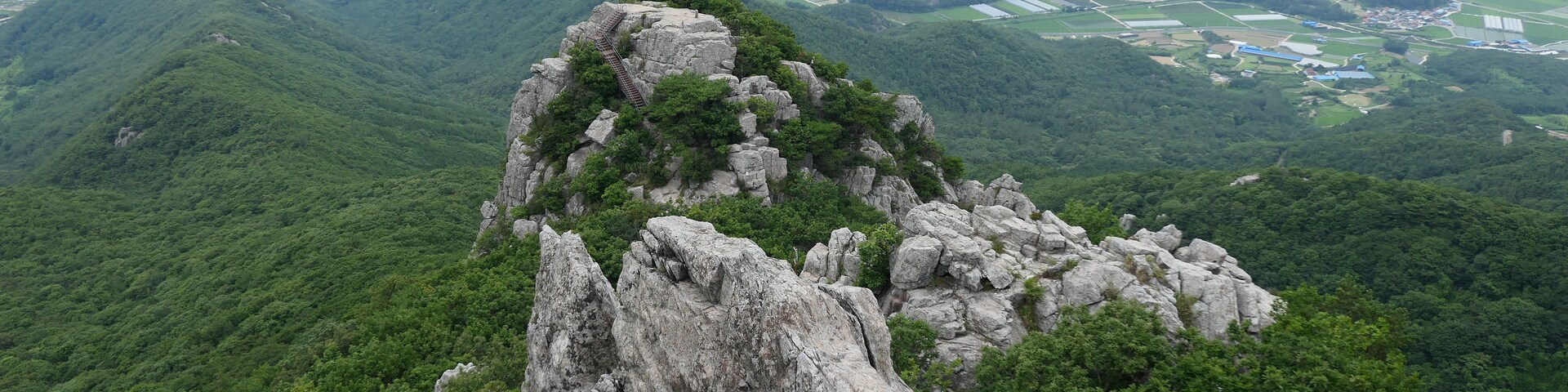Un paisaje montañoso con un pico rocoso destacado, una escalera que lleva a la cima y un valle con campos y un río a lo lejos.