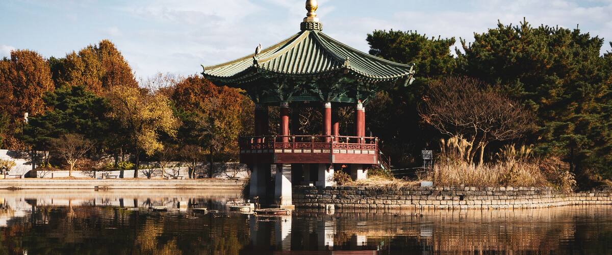 A traditional pavilion with a green roof and red pillars, surrounded by trees and a calm body of water.