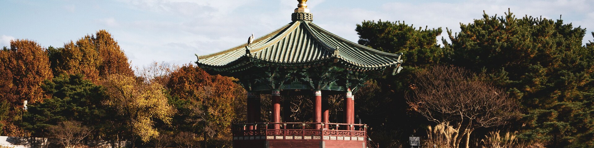 A traditional pavilion with a green roof and red pillars, surrounded by trees and a calm body of water.