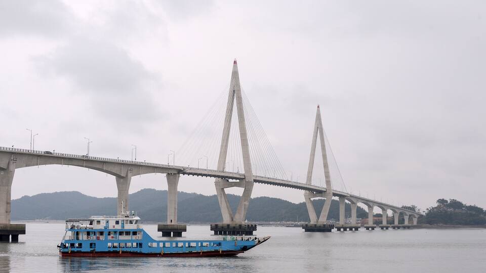 A blue ferry crossing a bridge with two tall pylons and a mountainous backdrop.