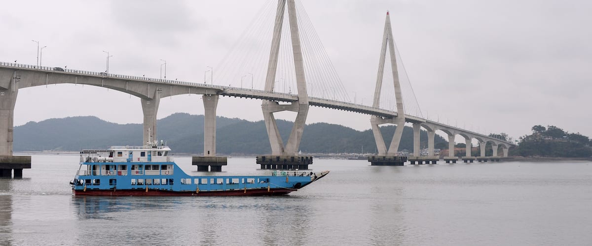 A blue ferry crossing a bridge with two tall pylons and a mountainous backdrop.