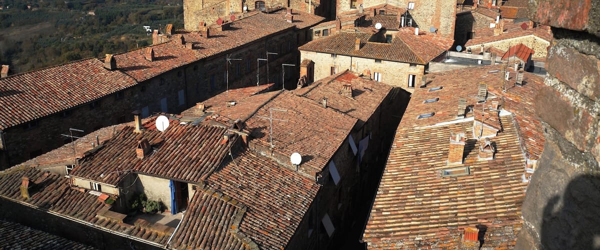 A historic town with terracotta-roofed buildings, a prominent church with a bell tower, and a hilly landscape in the background.
