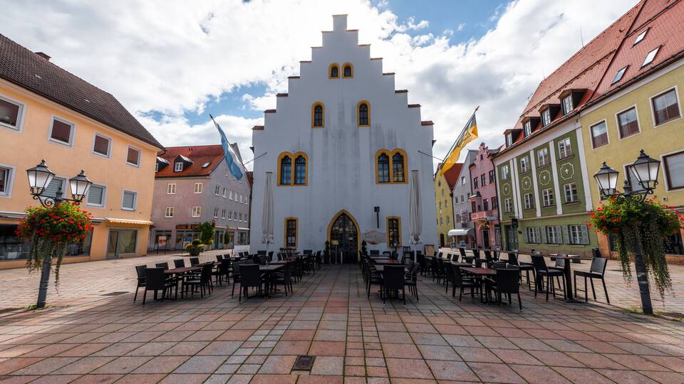A historic building with a steeply pitched roof and arched windows, surrounded by colorful buildings and outdoor seating.