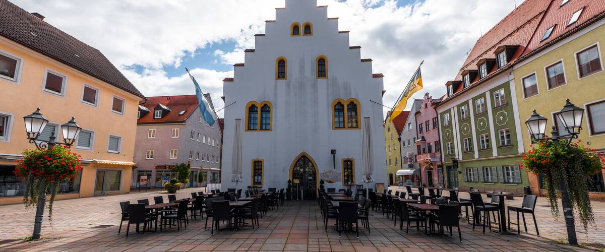 A historic building with a steeply pitched roof and arched windows, surrounded by colorful buildings and outdoor seating.