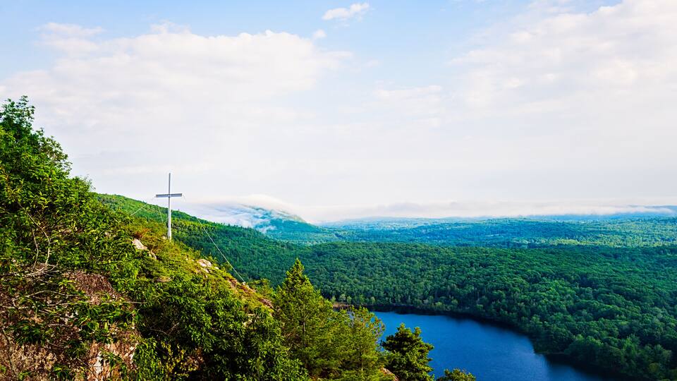 A cross on a hill overlooking a lake surrounded by dense forest.