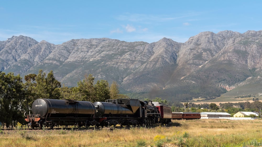 A steam locomotive with a tanker car in a rural area with mountains in the background.