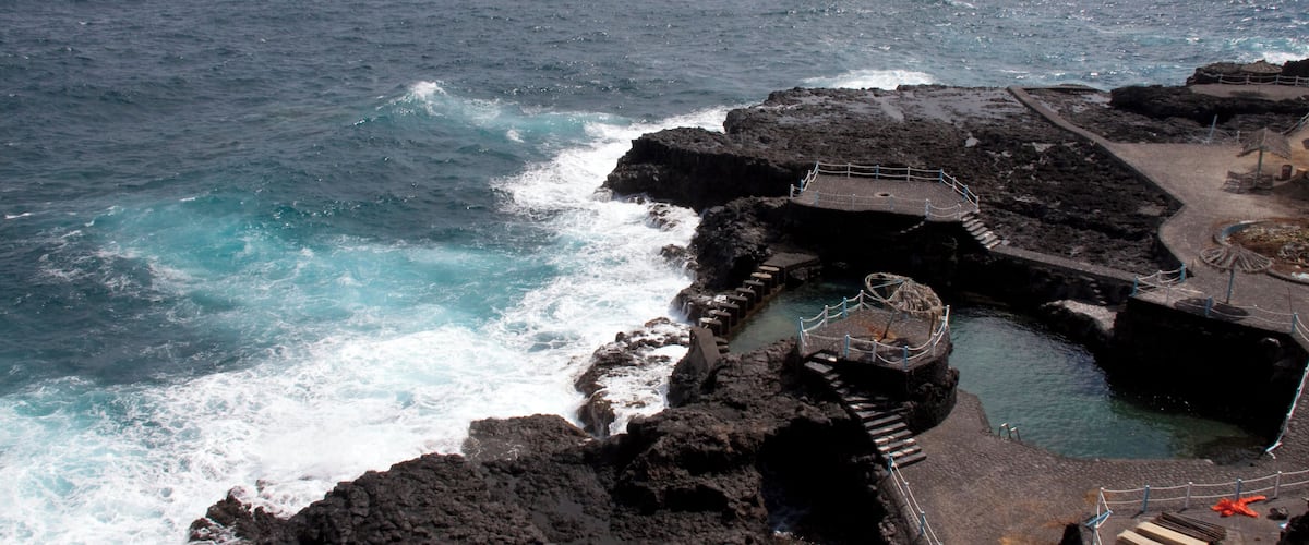 A rocky coastline with waves crashing against the shore, a small pool, and a staircase leading down to the water.