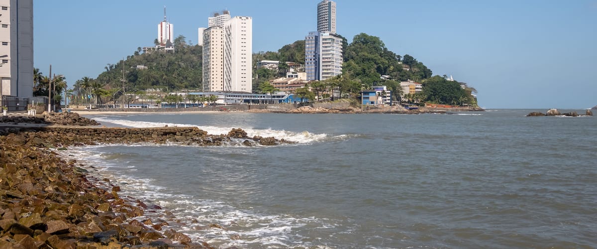 A coastal cityscape with high-rise buildings, a rocky shoreline, and a clear blue sky.