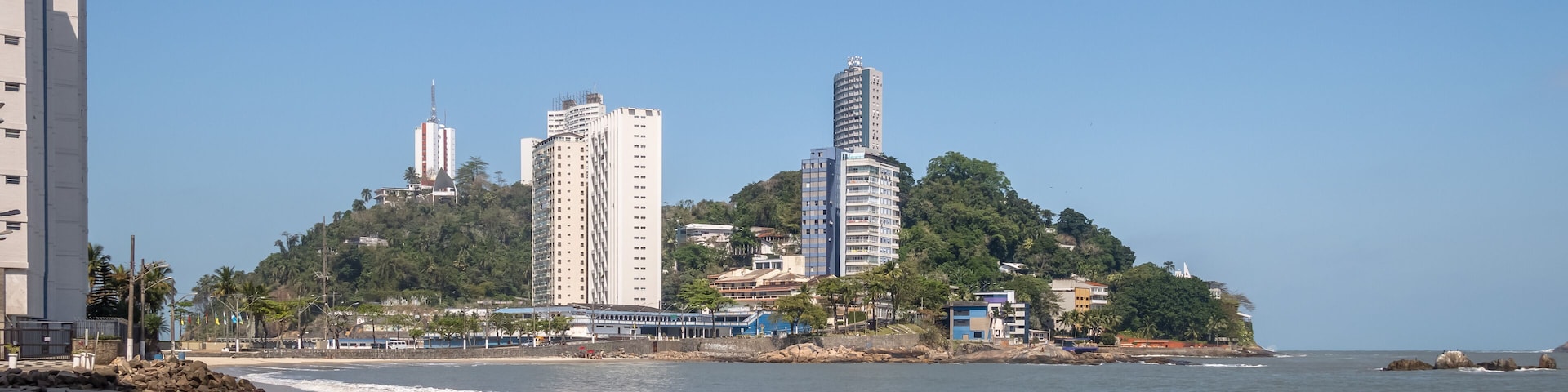 A coastal cityscape with high-rise buildings, a rocky shoreline, and a clear blue sky.