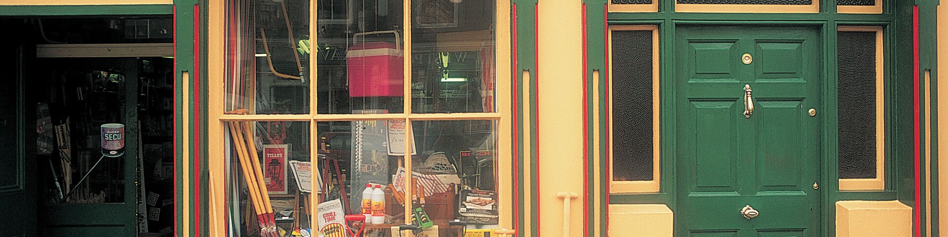 Fachada con una puerta verde y un escaparate que muestra herramientas de jardinería.