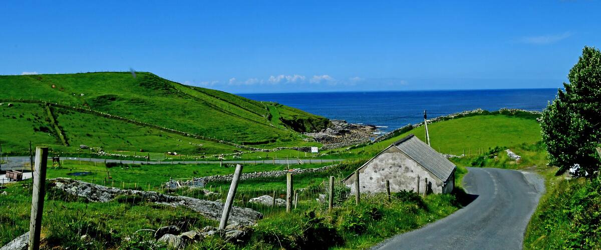 A rural landscape with a winding road, green hills, a stone wall, and a small building.