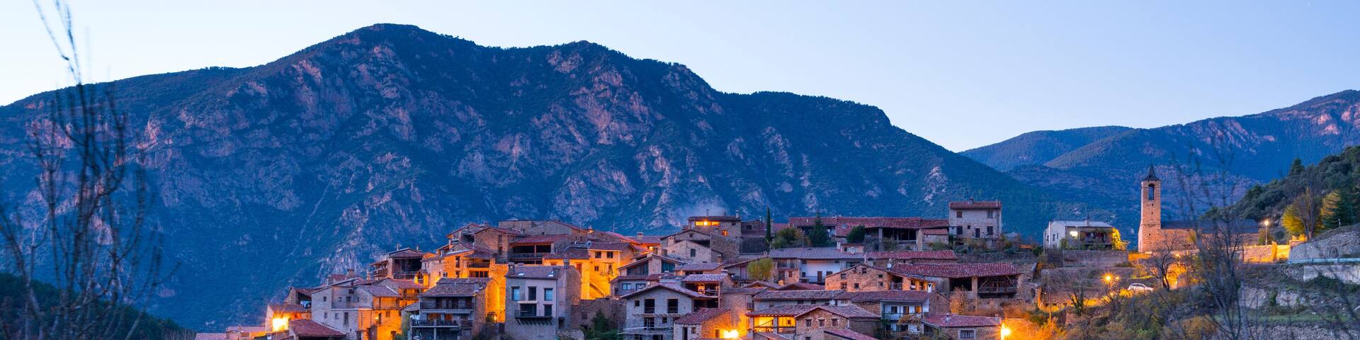 A hillside village with buildings illuminated at dusk, surrounded by mountains.