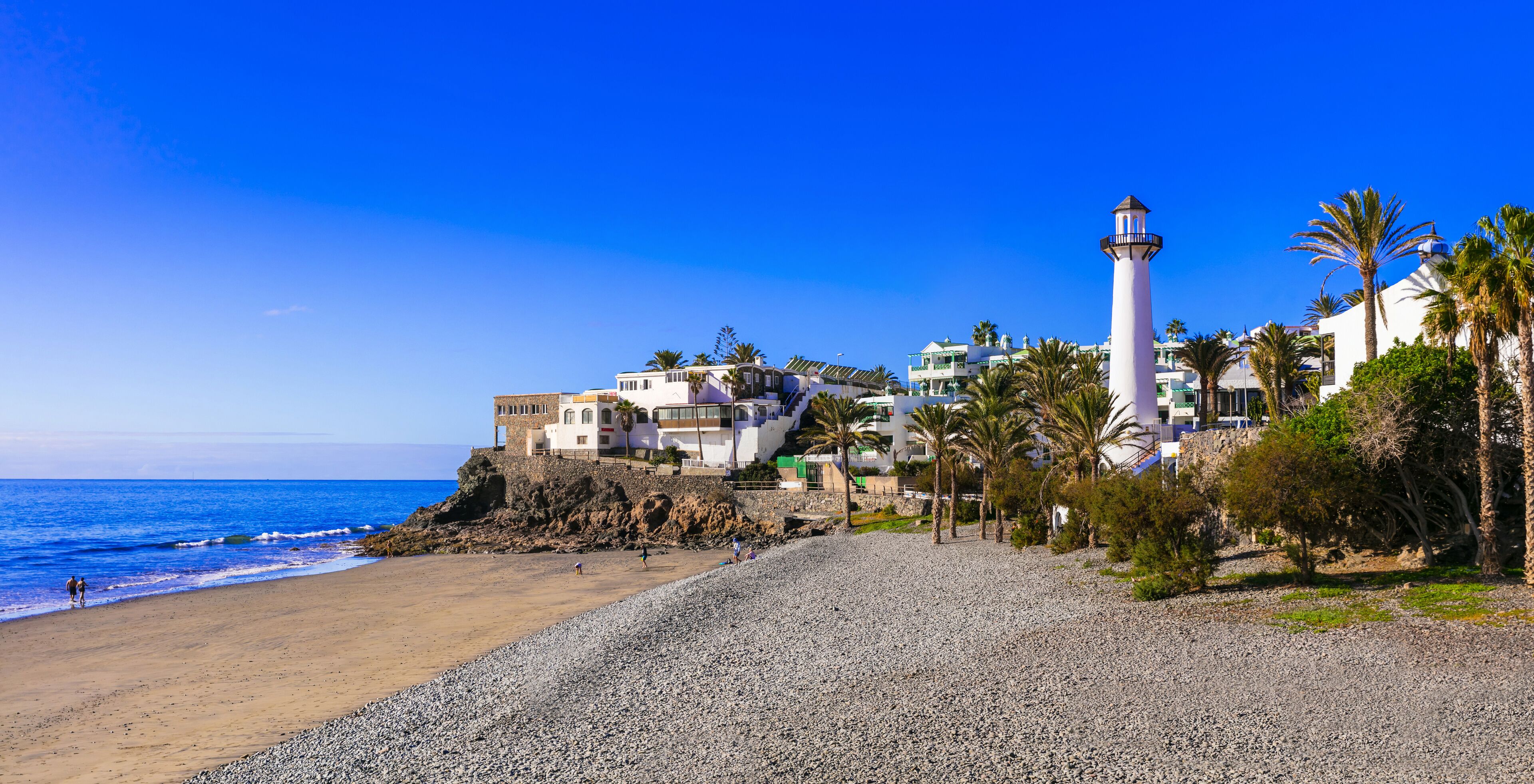A coastal scene with a beach, a lighthouse, and buildings on a rocky outcrop.