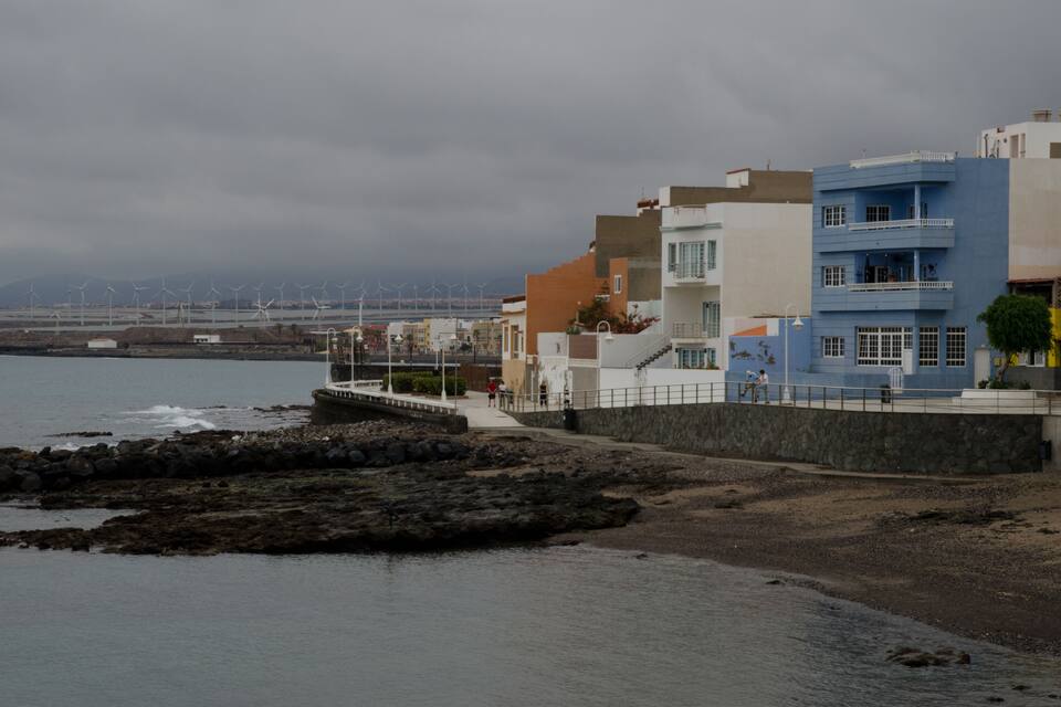 Une ville côtière avec des bâtiments qui bordent la plage, une plage rocheuse et des éoliennes visibles au loin.